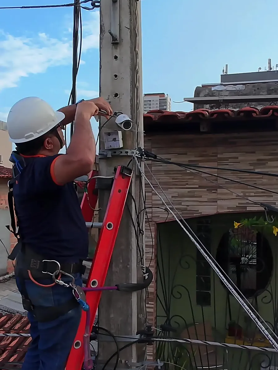 Técnico da Keleuthos com capacete e cinto de segurança instalando uma câmera de CFTV no alto de um poste